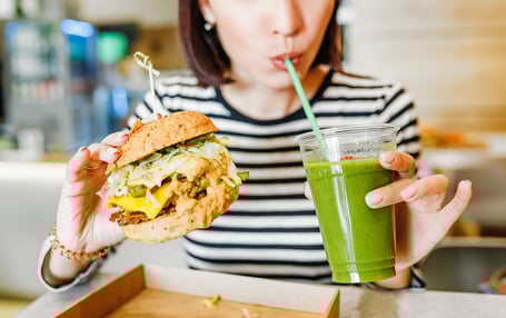 A woman drinking juice while holding a burger in another hand